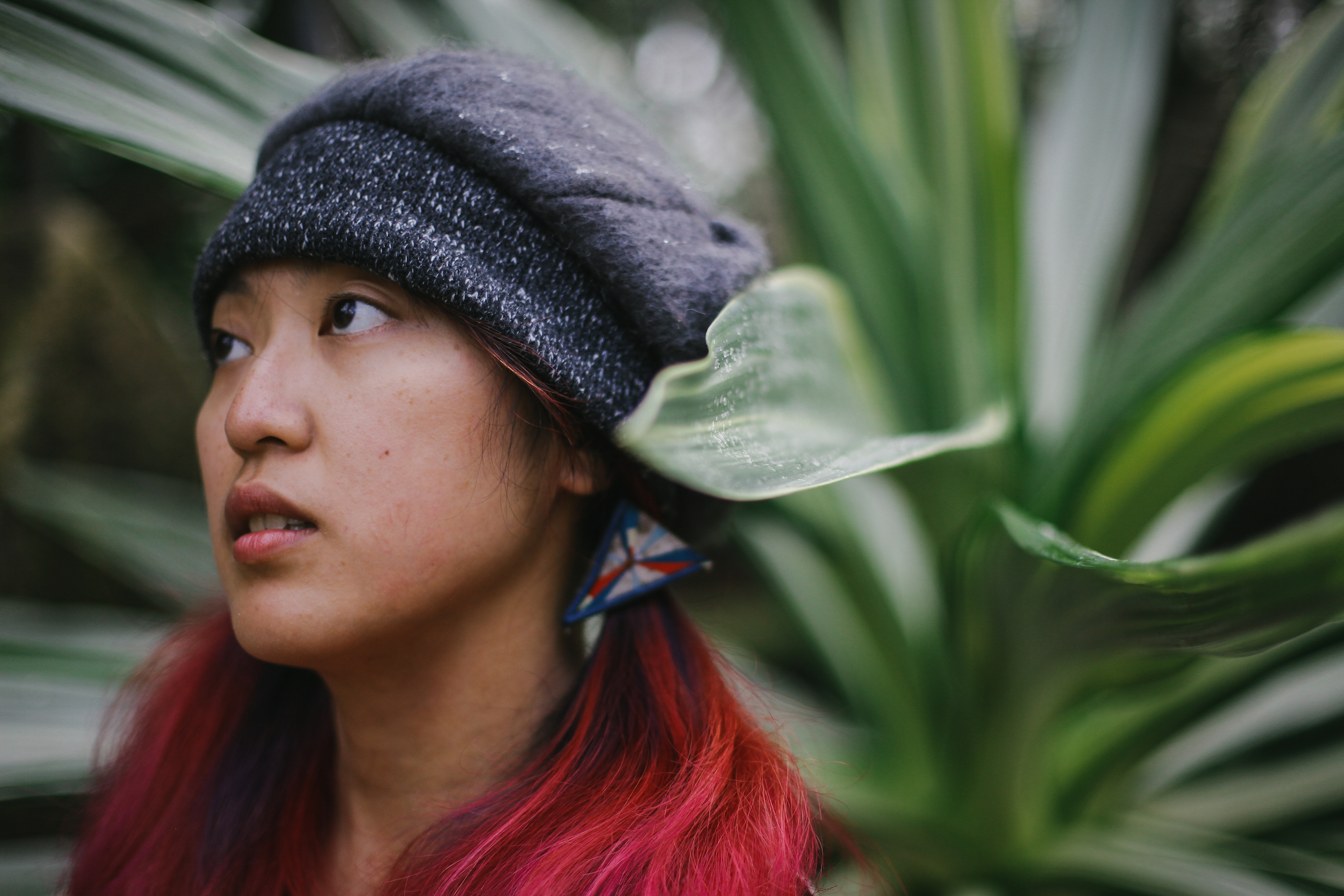 Young woman with long red hair and a dark blue cap on the left side looking in the distance to the left surrounded by a big green leafy plant